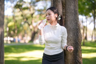 Yetişkin Asyalı kadın, canlı yeşil parktaki ağaca yaslanmış şişe suyla açık hava etkinliğini sürdürerek sulanır.