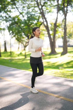 Enerjik Asyalı genç yetişkin kadın güneşli bir parkta açık havada koşmaktan hoşlanıyor. Odaklanma ve kararlılıkla fitness hedeflerine ulaşmak.