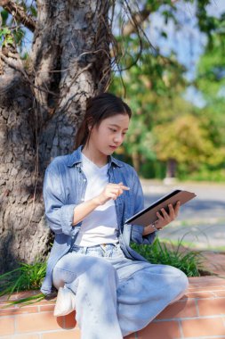 Yetişkin bir Asyalı kadın, hafta sonu güneş ışığı altında ağaçların yanındaki parkta sosyal medyayı izlemek için tablet kullanarak dijital yaşam tarzından keyif alıyor..