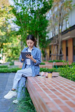 Genç Asyalı kadın üniversite parkında tabletle online çalışıyor. Eðitim ve öðrenim araþtýrmalarý için cep telefonu, kulaklık ve kahve fýrketi kullanan öðrenci.