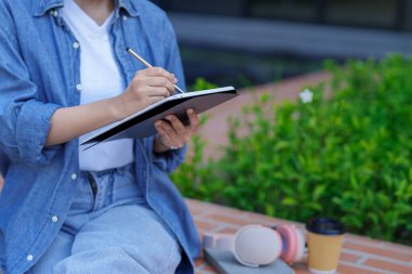 Asyalı yetişkin kadın, yeşil bitkilerin yanındaki bankta ders çalışmak için Stylus 'la tablet kullanıyor. Rahatlamak için sabah parkın tadını çıkarın..