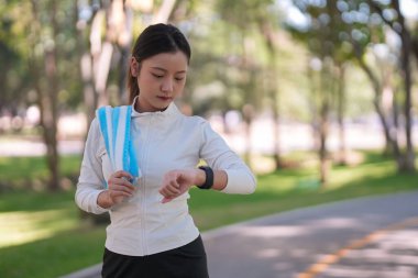 Genç Asyalı kadın, yeşil bir şehir parkında spor yaptıktan sonra akıllı saatleri kontrol ederek aktif yaşam tarzını gözlemliyor.