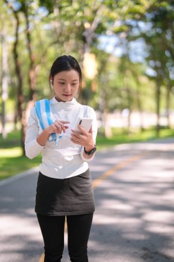 Enerjik Asyalı kadın, yeşil parkta koştuktan sonra mobil cihazları kontrol ediyor. Sağlık ve aktif yaşam tarzı için modern teknolojiden hoşlanıyor.