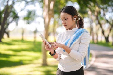 Yetişkin Asyalı kadın park molasından hoşlanıyor. Telefonları dinleyip açık havada spor yapmak ve refah içinde yaşamak için dinliyor..