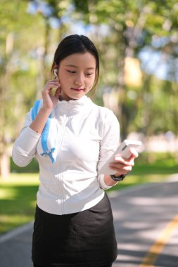 Asyalı yetişkin kadın, kablosuz kulaklığı açık havada elinde telefonla ayarlıyor. Aktif yaşam tarzına odaklanın güzel park ortamları.