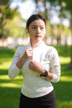 Asyalı kadın atletizm ceketini canlı bir parka yerleştiriyor ve modern teknoloji ile aktif yaşam tarzı sağlığı ve refahı vurguluyor.