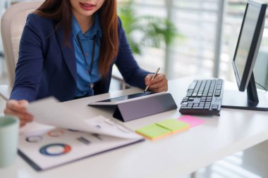 Asyalı yetişkin kadın ofiste çalışıyor. Bir tablet Stylus kullanıyor, mali verileri ve iş raporlarını gözden geçiriyor. Modern teknolojiye odaklan..
