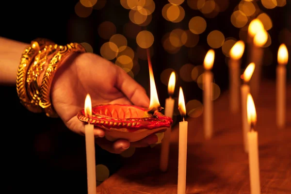 Indian housewife or bride woman wearing traditional gold jewelry, lighting candles with a clay diya or oil lamp in one hand at a temple on Diwali night. Religious ritual.