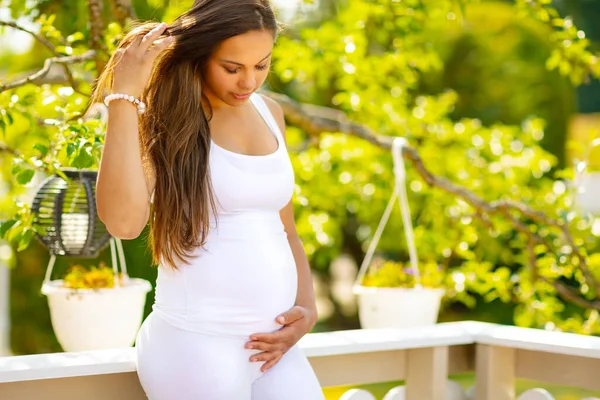Счастливая беременная женщина, стоящая в саду, держа руки на животе — стоковое фото