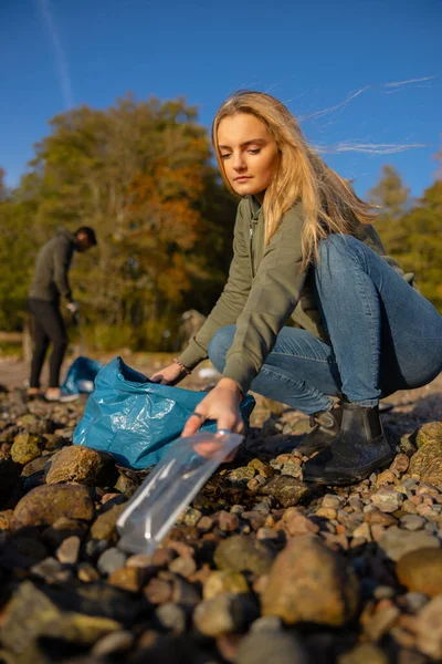 Serious young woman picking up plastic at rocky beach — Stock Photo, Image