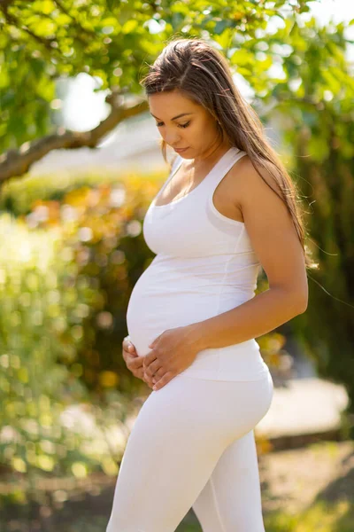 Těhotná žena stojí v zahradě a dívá se na své břicho — Stock fotografie