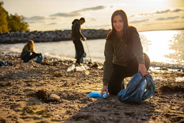 Mujer joven enfocada limpiando la playa con un equipo de voluntarios durante el atardecer — Foto de Stock