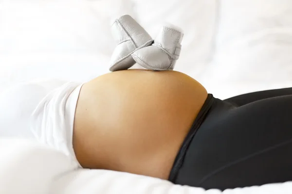 Femme enceinte au lit avec des chaussures de bébé sur le ventre — Photo