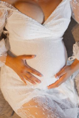 Görüntü, anne adayının karnını nazikçe tutması gibi huzurlu bir anı tasvir ediyor. Beyaz dantelli elbisesi saflığı ve yeni bir hayatın beklentisini sembolize ediyor, neşe ve annelik bağlılığını ifade ediyor..