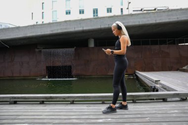 Aktif giysi giyen genç bir kadın şehirde yürürken ve egzersiz yaparken müzik dinliyor..