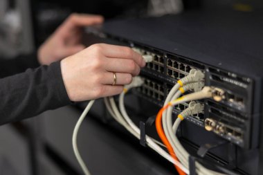 30 'lu yaşlarda beyaz bir kadın veri tabanında çalışıyor, bir sürü şebeke anahtarıyla uğraşıyor. Kabloları takıyor ve güvenli bağlantıyı sağlamak için bakım, teşhis ve sorun giderme işlerini yapıyor. Sahne teknik beceri, odaklanma ve