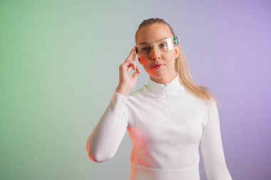 Metalik yansıtıcı gözlük takan ve beyaz giysili bir kadın çok renkli bir arka plana karşı poz veriyor. Jesti, fütüristik bir temayı vurgulayarak, düşünmeyi veya sanal elementlerle etkileşimi öneriyor. Yumuşak ışıklandırma ve renk