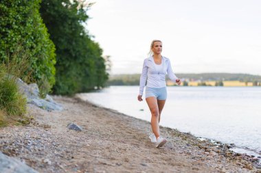 A woman walks along a tranquil lakeshore wearing sports attire, portraying wellness and mindfulness.