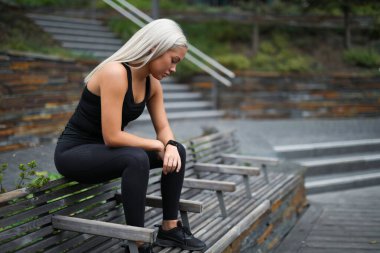 Aktif kıyafetli genç bir kadın, sakin bir şehir parkında, ahşap bir bankta oturmuş antrenman seansından sonra düşünüp taşınıyor. Arkaplan özellikleri yeşillik ve yapılandırılmış ahşap elementleri düzenledi.