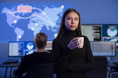 Ekranlarla aydınlatılmış bir teknoloji kontrol odasında, kadın bir siber güvenlik uzmanı gerçek zamanlı veri tabanlarını analiz ederken kahve içer. Ayarlama takım çalışmasını ve tehditte kullanılan gelişmiş izleme sistemlerini vurgular