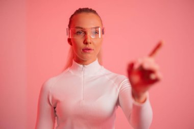 Minimalist beyaz giysili bir kadın fütürist gözlük takıyor ve elini bilerek hareket ettiriyor. Pembe arka plan ve yumuşak ışıklandırma yenilik ve yaratıcılığı vurgulayan modern ve teknolojik bir atmosfer yaratıyor. Bu resim,