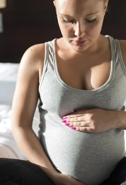 Pregnant woman sits on bed with her hand at belly