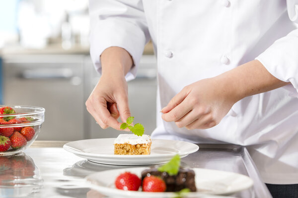Close-up of a professional female chef who decorates dessert cake with strawberry and lemon leaf. Large industry kitchen.