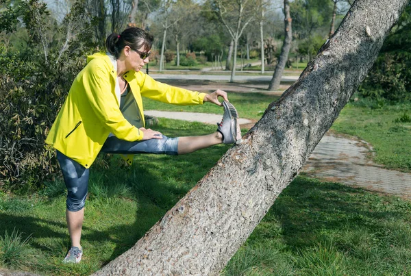 FEMALE RUNNER IN YELLOW WINDBREAKER STRETCHING WHILE LEANING AGAINST A TREE