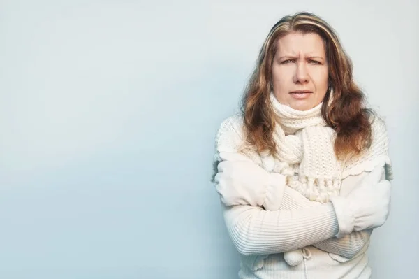 A middle-aged woman shows that she\'s frozen. She is dressed in a white warm sweater and white mittens on a blue background. The woman hugs herself to warm up