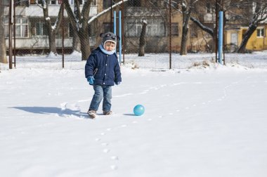 Küçük çocuk kışın sonunda karda bir topla koşuyor. Sıcak kış kıyafetleri ve köpek şeklinde bir şapkası var.