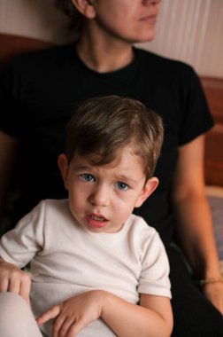 Küçük çocuk annesinin dizlerinde ağlıyor. Çocuğun üzgün, ağlayan bir yüzü var, gözlerinden yaşlar akıyor. Dikey