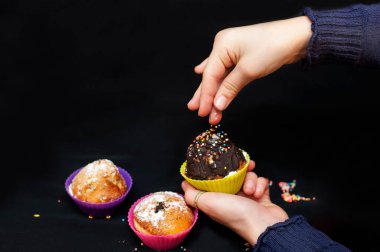 Genç kız şekerleme serpme renkli şekerleme siyah arka planda çikolatalı mafinleri süsler. Uzayı kopyala Yatay