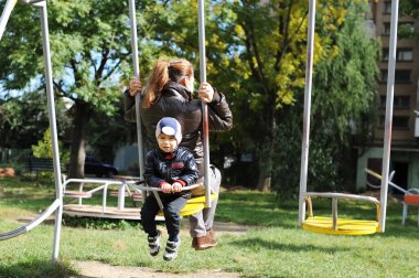 Anne ve küçük oğlu bahar ya da sonbaharda salıncağa biniyor. Çeşitli insanlar - yetişkin bir kadın ve küçük bir çocuk, onun oğlu