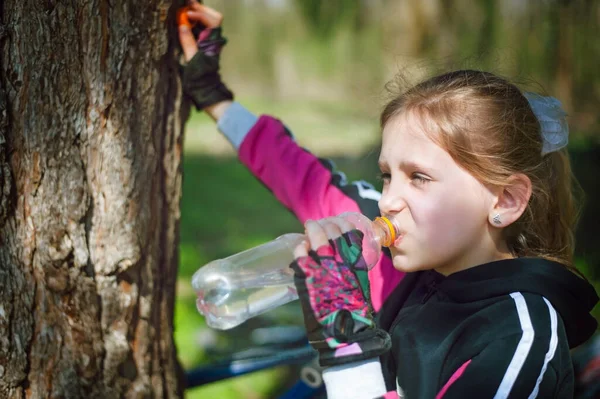 A girl while biking is resting at the tree and drinking clean water from the bottle. She is dressed in a sports suit and gloves for a bicycle. She has a happy face