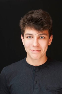 Beautiful teenager with courageous cheerful face and dark hair smiles on black background. Vertical. Portrait of happy beautiful guy