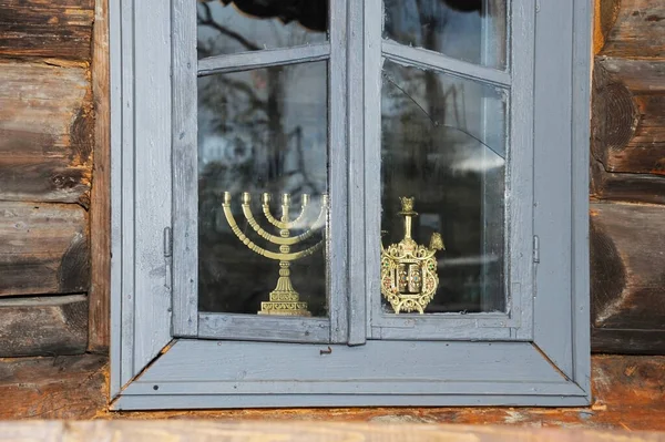 An old window with broken glass in an old wooden house. On the window there are symbols of the Jewish people - candlesticks of the Menorah. Vertical