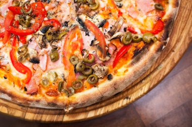 Domatesli, peynirli, yeşil zeytinli, mantarlı ve jambonlu büyük bir pizza ahşap bir pizza standında. Kapatın. Üst görünüm