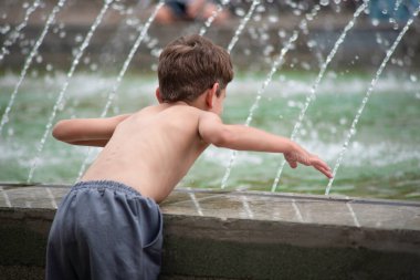 Küçük çocuk yazın sıcaktır ve gömleğini çıkardı ve ısıya daha kolay dayanmak için çeşmeye suyla atlamak istiyor gibi görünüyor. Küçük bir çocuk çeşme suyuna atlar.