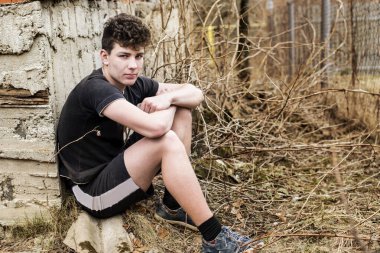 Üzgün yüzlü, şort ve tişört giymiş bir genç ormanda eski bir evin yanında oturuyor ve kaybolduğu için eve nasıl gitmesi gerektiğini düşünüyor.