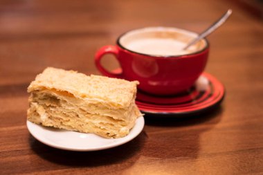Napolyon adındaki bir puf böreğinin yanında, ahşap bir masanın üzerinde büyük kırmızı bir fincanda kapuçino. Lezzetli bir aperatif.