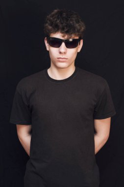 Siyah gömlekli ve siyah arka planda güneş gözlüklü ergenlik çağındaki bir çocuk. Çocuğun ciddi bir yüzü var. Dikey
