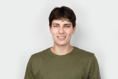 Genç bir çocuk, hala mutlu bir genç, açık arkaplanda gülümseyen, haki rengi bir tişört giyen, koyu saçlı ve beyaz dişli. Çocuğun keyfi yerinde ve depresyonda değil, reklam verin.