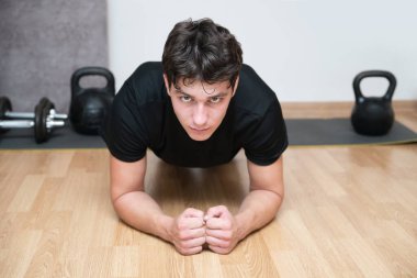 Genç bir adam, fiziksel egzersiz yapıyor, tahta pozisyonda duruyor, kolay biri olmadığı açık, yüzü kırmızı. Adam statik egzersizleri, kalas egzersizlerinin çeşitlerini seviyor.