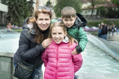 Bir çocuk kız kardeşiyle ve annesiyle fıskiyenin yanındaki şehirde dinlenip fotoğraf çekiyor. Anne ve çocuklar birlikte eğleniyorlar.