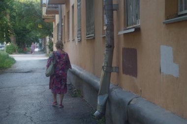 Şehir hayatının bir sahnesinin fotoğrafı. Renkli sabahlık giymiş gri saçlı yaşlı bir kadın eski ağaçların gölgesinde, eski bir evin yanında yürüyor..