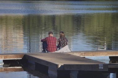 Genç bir çift bir göletin kıyısında dinleniyor. Su yansımalar ve eğimlerle dalgalanır. Güneşli bir akşam. Su sporları için bir iskele..