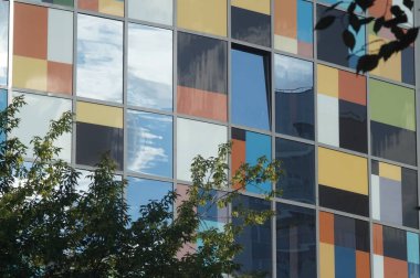 Photo of a part of the facade of a new building. Reflective glass and colored plastic. Clouds are reflected in the facade. Rich colors.