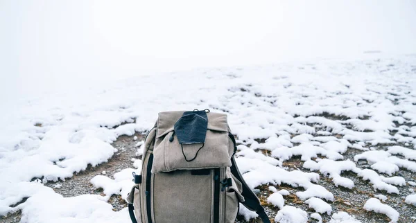 Travel backpack with a medical face mask on a snowy background. Concept of traveling in winter 2020 with the covid-19 pandemic.
