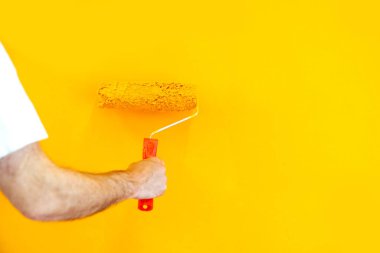 Hand of an anonymous painter holding a roller painting a orange wall at work, closeup view. Offering professional painting services. Isolated on big empty space.