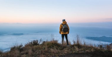 Genç bir gezgin, güzel bir gün doğumunda sisli bir denizle kaplı araziyi gözlemliyor. Dağın tepesindeki maceraperest gezginin dikiz görüntüsü. Seyahat kavramı, doğa ve özgürlük..